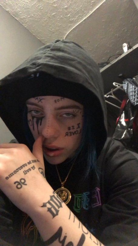 Billie covered in face tattoos, wearing a gold chain and black hoodie. tattoos all over her face and hands like lil Xan